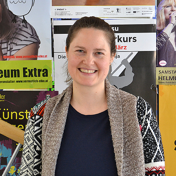 Portrait einer Frau mit Pferdeschwanz, die lächelnd vor einer Plakatwand steht