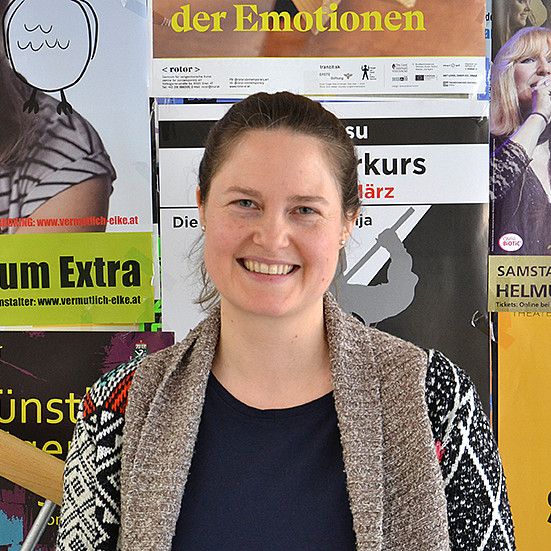 Portrait einer Frau mit Pferdeschwanz, die lächelnd vor einer Plakatwand steht