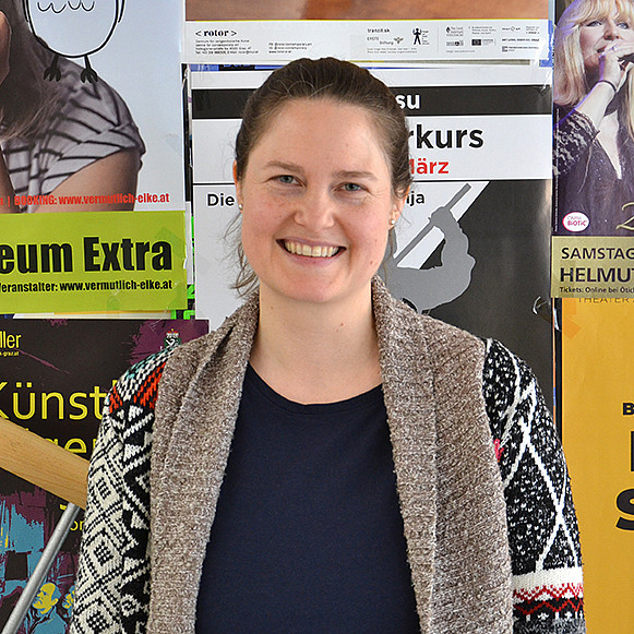 Portrait einer Frau mit Pferdeschwanz, die lächelnd vor einer Plakatwand steht