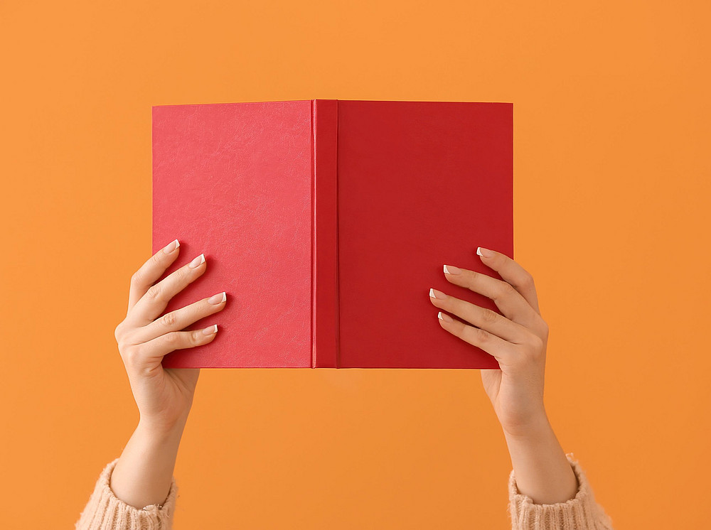 Hands holding up a book with a red cover against an orange background ©Pixel-Shot - stock.adobe.com
