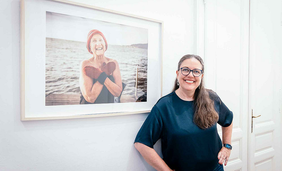 eine lächelnde Frau steht vor einem Bild, auf der eine ältere Frau zu sehen ist