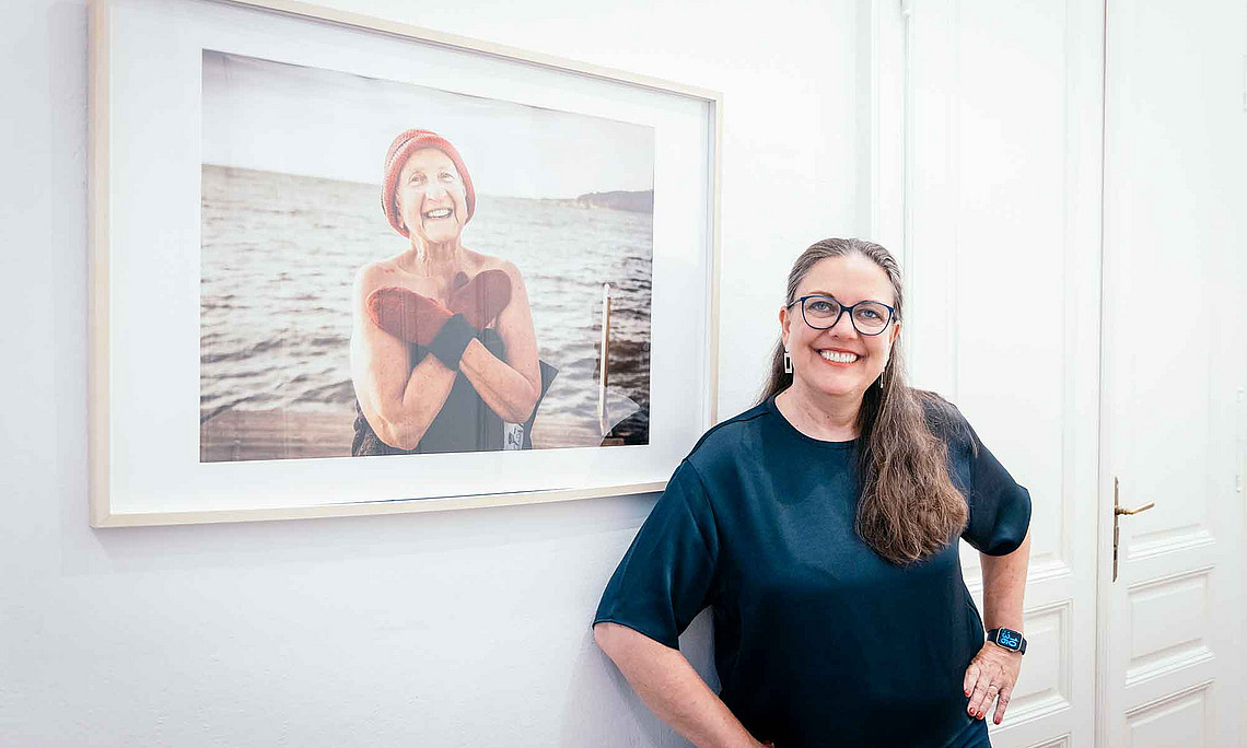 eine lächelnde Frau steht vor einem Bild, auf der eine ältere Frau zu sehen ist