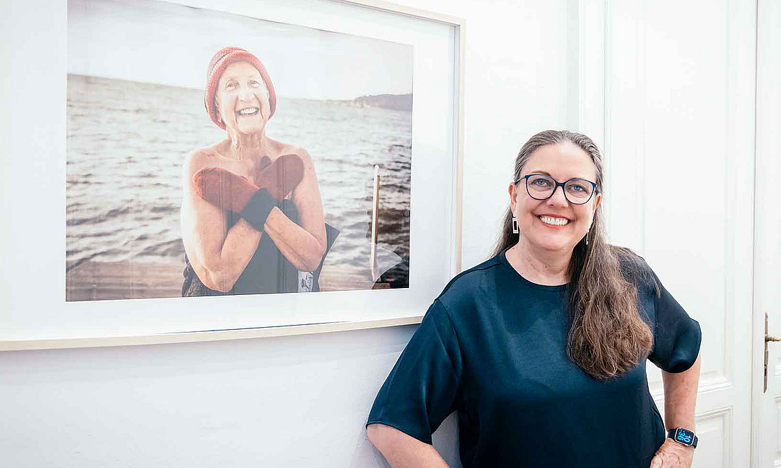 A smiling woman is standing in front of a picture showing an elderly woman