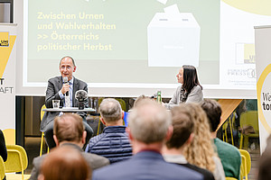 A panel discussion with a leading political analyst and a journalist in an office conference room. The two people are sitting at their desks and speaking to the audience. There are subtle yellow highlights in the background, emphasising important elements such as banners, seating arrangements and the composition of the crowd.