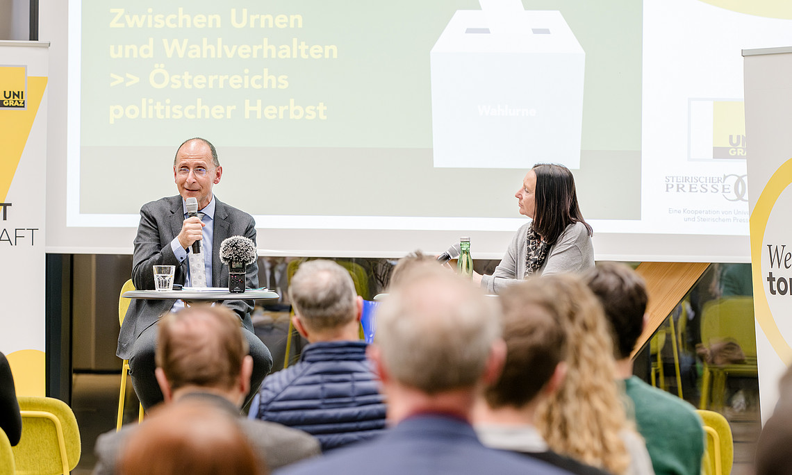 A panel discussion with a leading political analyst and a journalist in an office conference room. The two people are sitting at their desks and speaking to the audience. There are subtle yellow highlights in the background, emphasising important elements such as banners, seating arrangements and the composition of the crowd.