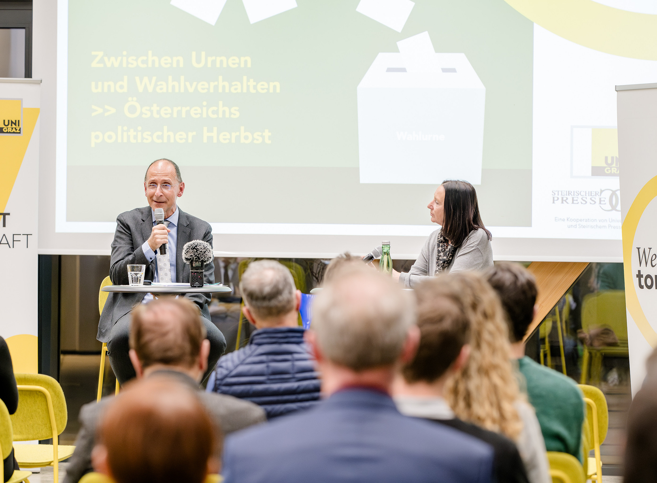 Eine Podiumsdiskussion mit einem führenden politischen Analysten und einer Journalistin in einem Bürokonferenzraum. Die beiden Personen sitzen an ihren Schreibtischen und sprechen zum Publikum. Im Hintergrund sind dezente gelbe Akzente zu sehen, die wichtige Elemente wie Banner, Sitzordnung und Zusammensetzung der Menge hervorheben. ©MICHAELA PFLEGER