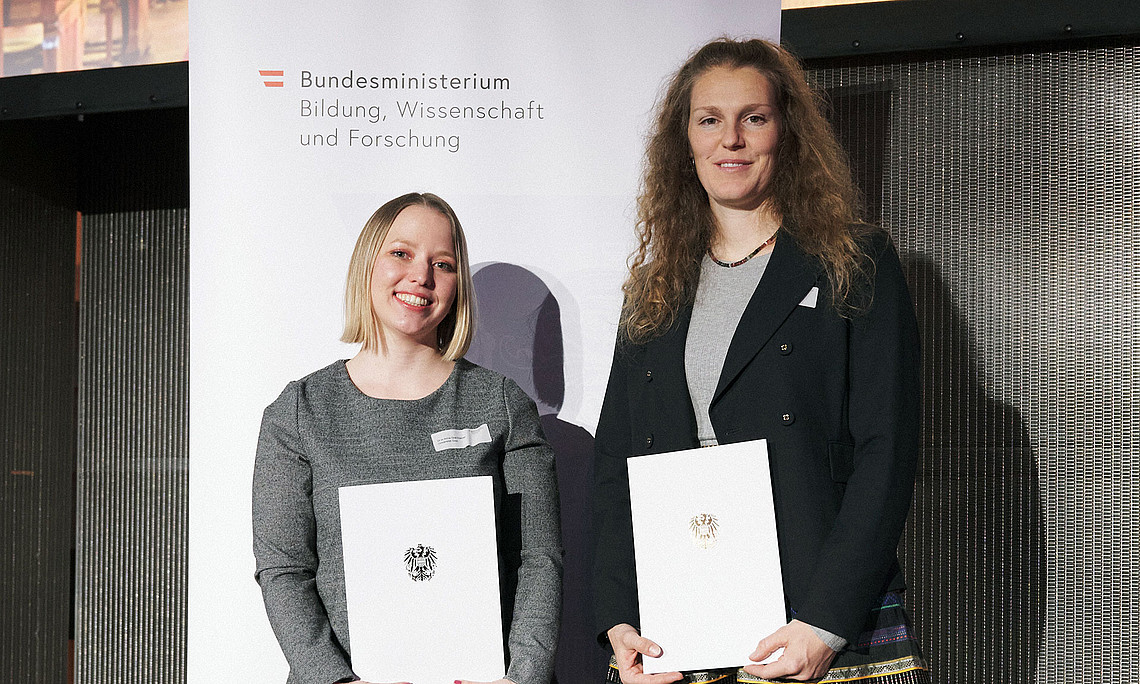 Anna Graf-Steiner und Marina Maren Reiner mit Urkunden vor einem Roll-up des BMBWF