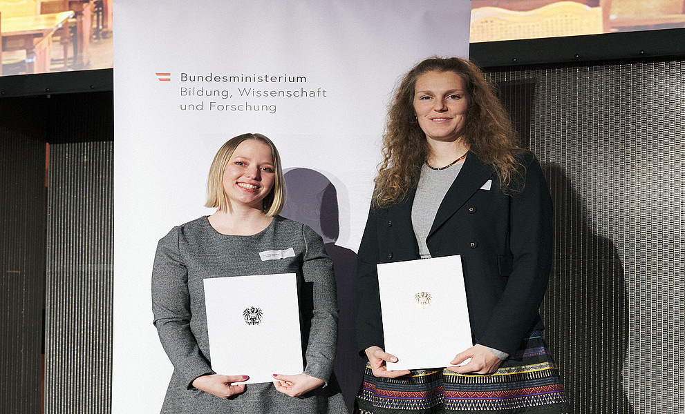 Anna Graf-Steiner und Marina Maren Reiner mit Urkunden vor einem Roll-up des BMBWF