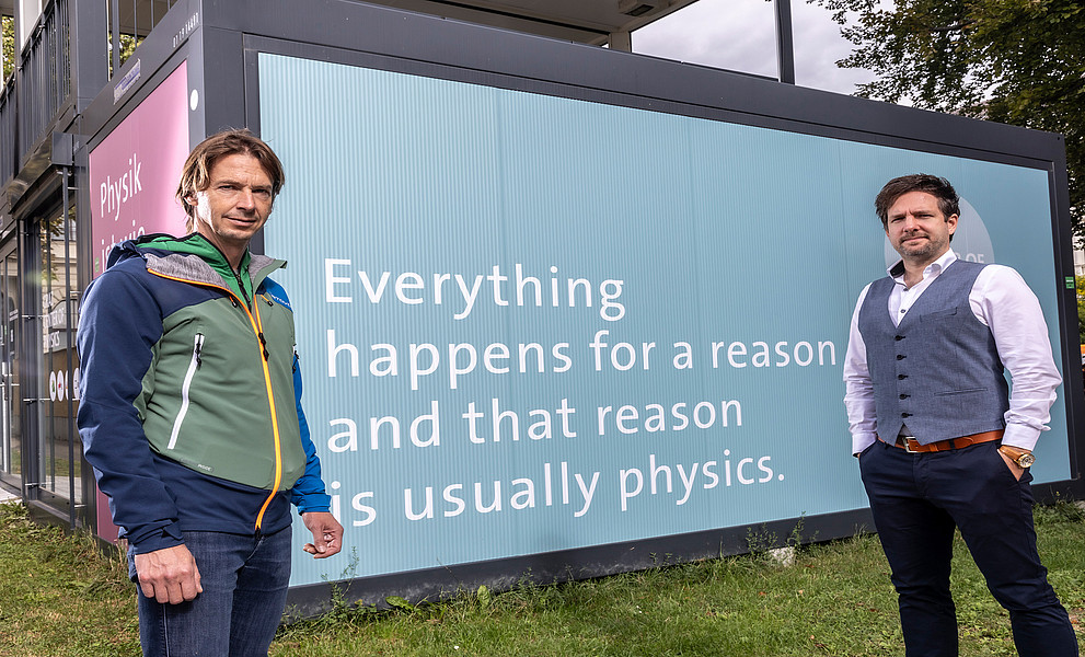 Alexander Bergmann und Peter Banzer vor dem Info-Point zum entstehenden Graz Center of Physics mit der Aufschrift Everything happens for a reason and that reason is usually physics". 