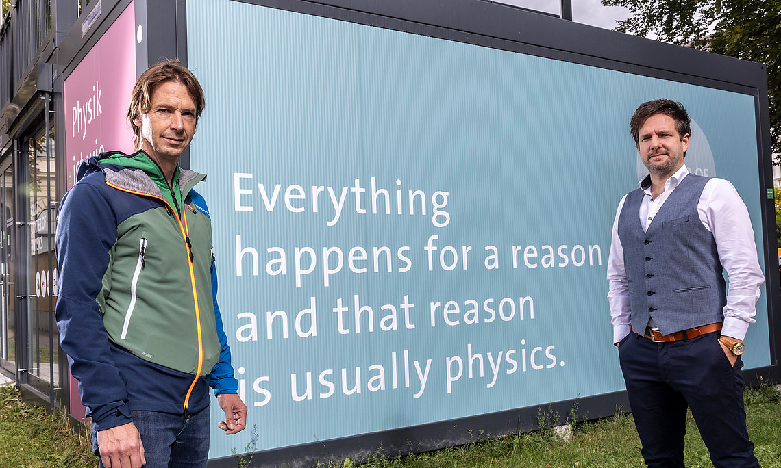 Alexander Bergmann and Peter Banzer in front of the info point for the emerging Graz Center of Physics with the inscription "Everything happens for a reason and that reason is usually physics". 