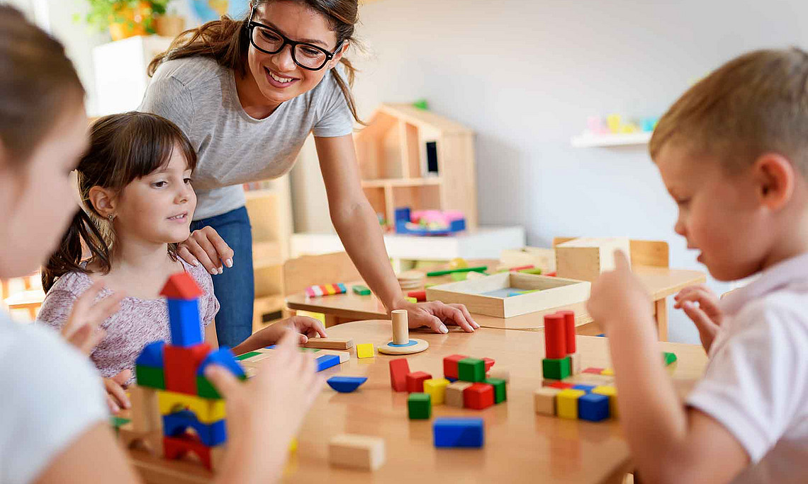 An adult wearing glasses is leaning over a children’s table; three children are sitting in front of it, building with colourful wooden blocks 
