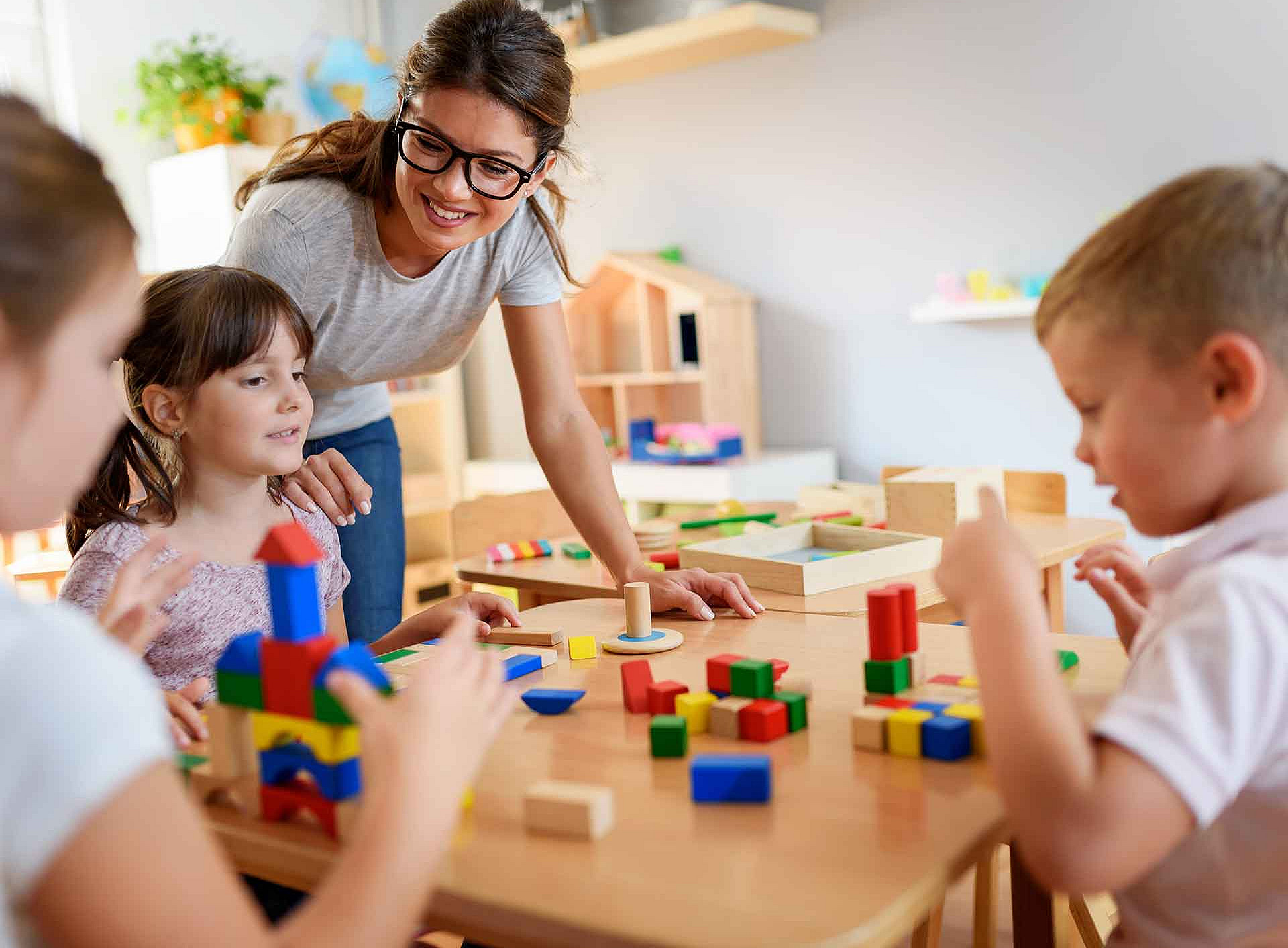 An adult wearing glasses is leaning over a children’s table; three children are sitting in front of it, building with colourful wooden blocks ©nemanfoto@gmail.com