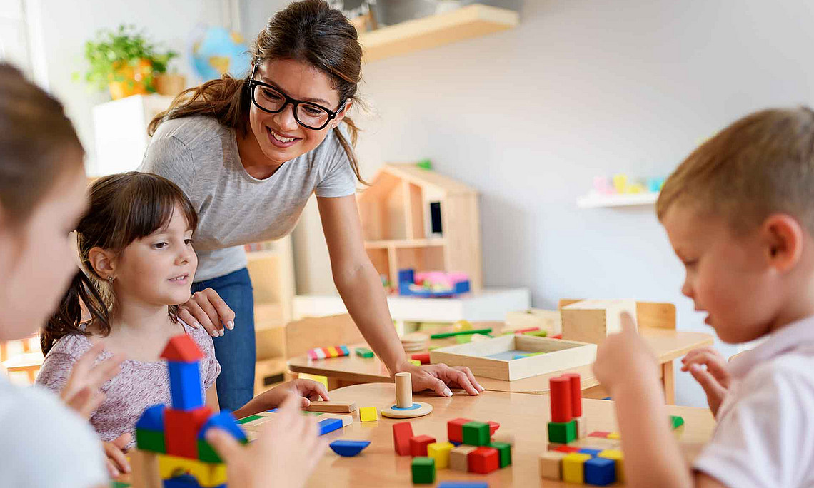 Erwachsene mit Brille beugt sich über Kindertisch, drei Kinder sitzen davor und bauen mit bunten Holzklötzen