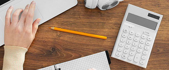 Schmuckbild Eine Hand, ein Laptop, ein Bleistift und ein Taschenrechner auf einem Schreibtisch.