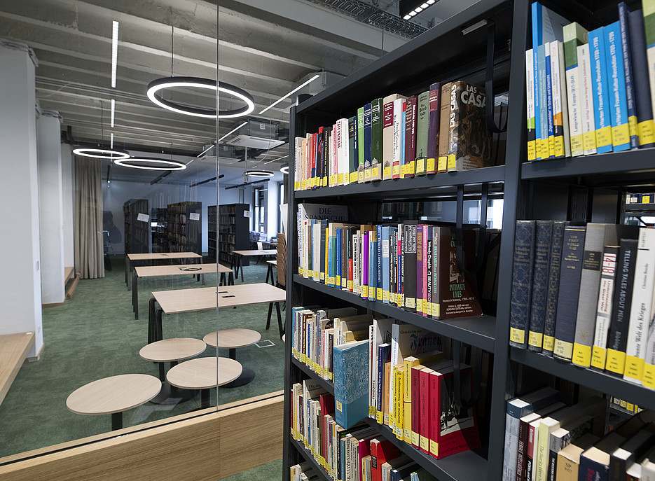 „Blick vom Rand eines Bücherregals mit bunten Buchrücken in einen großzügigen Bibliotheks- und Lernbereich mit mehreren Arbeitstischen, runden niedrigen Beistelltischen, grünem Teppichboden und auffälligen ringförmigen Deckenleuchten.“ ©Uni Graz/Tzivanopoulos