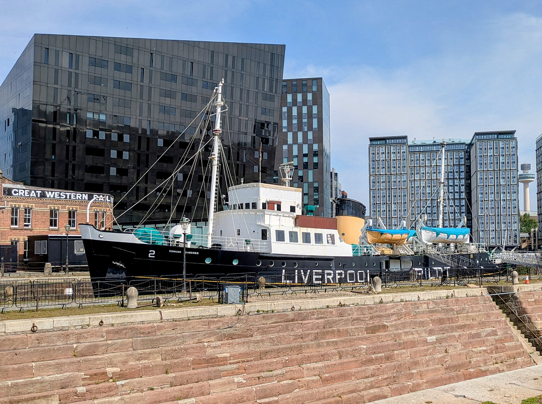 Das Foto zeigt ein historisches Schiff mit der Aufschrift „Liverpool“, das neben modernen Gebäuden in der Nähe einer Uferpromenade vor Anker liegt. Die industrielle Architektur steht im Kontrast zum traditionellen Design des Schiffes. Die Szene spielt sich unter einem klaren Himmel ab. ©Daniella Shaw