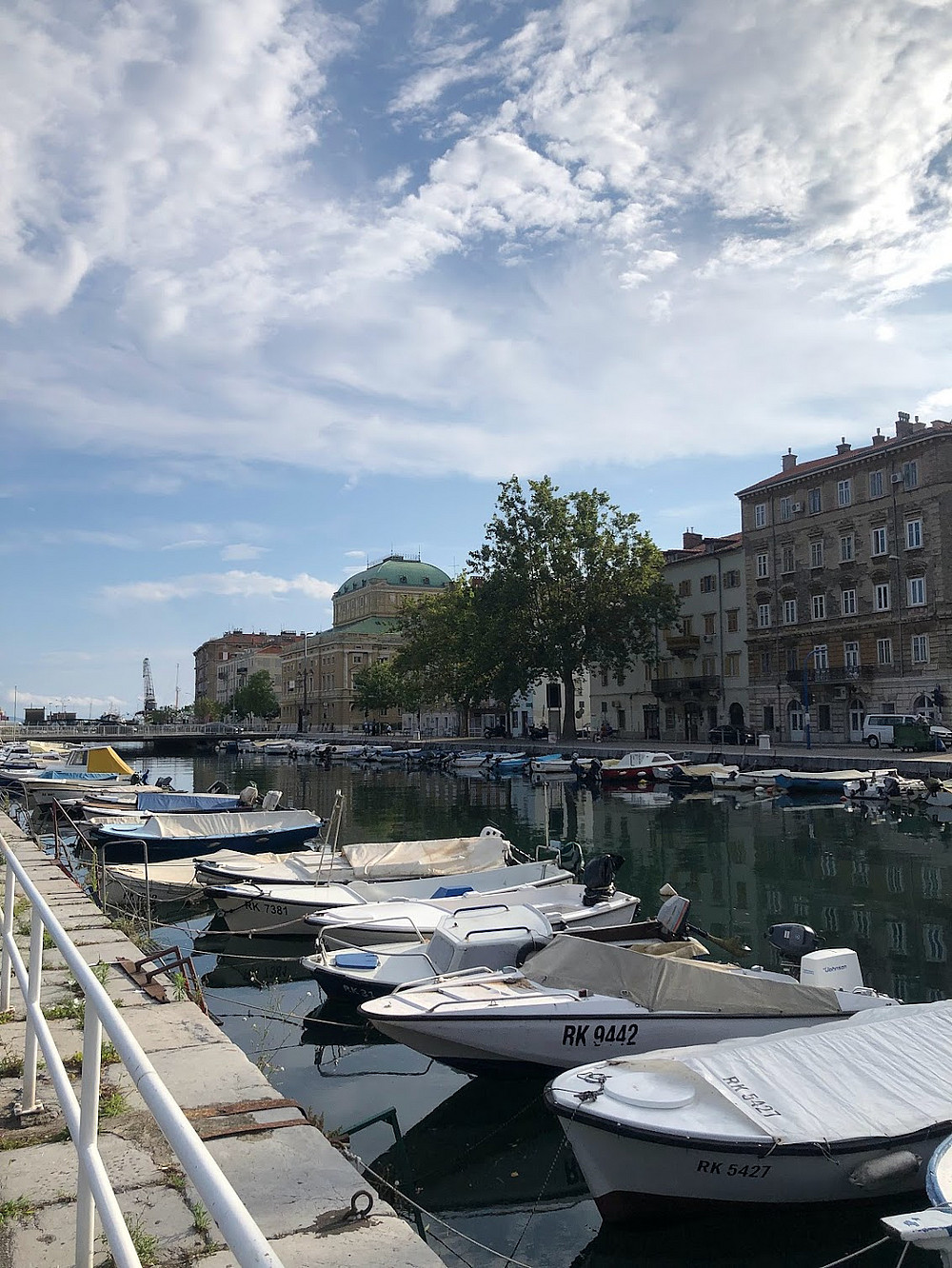 Boats docked along a calm waterfront, bordered by historic buildings under a partly cloudy sky. The scene conveys a serene, picturesque ambiance. ©Milka Car