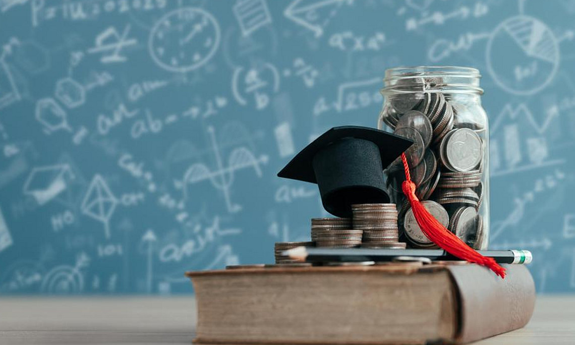 A doctoral cap and a pile of coins are lying on a book. In the background, a blackboard covered in writing can be seen. 