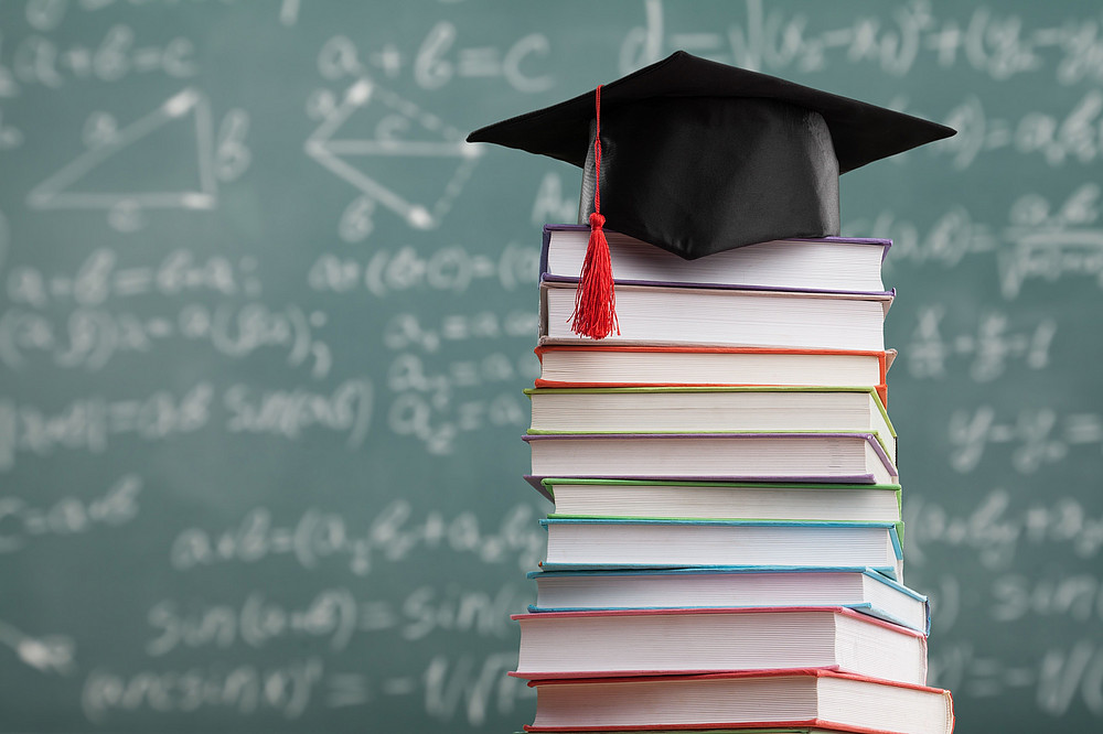 A pile of books in front of a blackboard with a doctor's hat on it ©BillionPhotos.com - stock.adobe