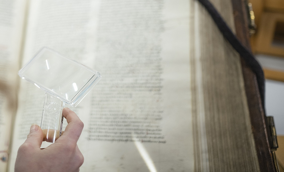 Eine Hand hält eine Lupe und vergrößert eine mittelalterliche Handschrift