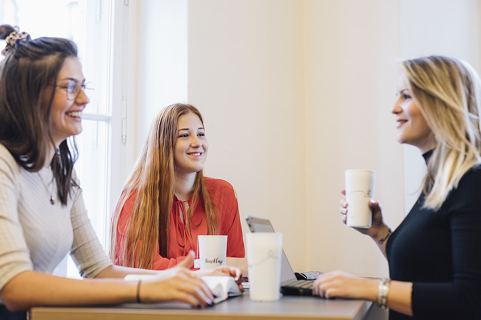 Drei Studierende sitzen mit To-Go-Heißgetränken gemeinsam im Innenbereich an einem Tisch und unterhalten sich fröhlich.
