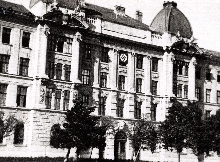 Historisches, mehrstöckiges Gebäude mit Kuppeldach, mittig ein Hakenkreuz-Symbol an der Fassade, davor Bäume. 