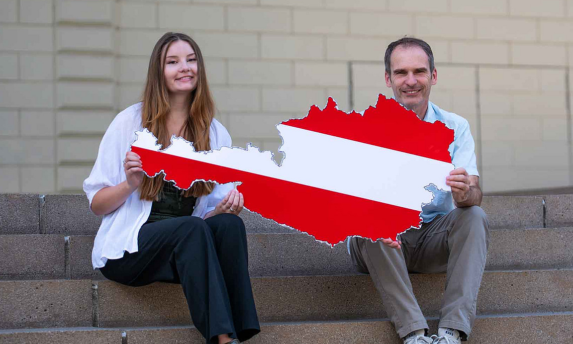 Ein Mann und eine jüngere Frau sitzen auf einer Treppe und halten gemeinsam eine große rot-weiß-rote Karte von Österreich vor sich.