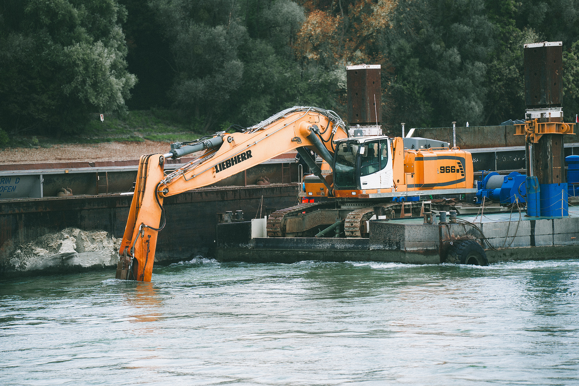 Orange excavator on a floating platform dredging a river, with its arm extended into the water and trees in the background. ©Manuel Madaini