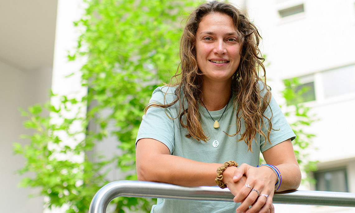 Florina Schalamon, mit den Unterarmen auf eine Reling gestützt, vor einem weißen Gebäude und grünem Baum