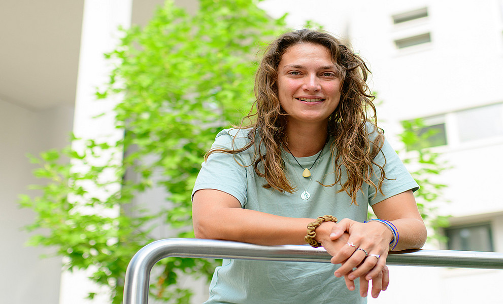 Florina Schalamon, mit den Unterarmen auf eine Reling gestützt, vor einem weißen Gebäude und grünem Baum