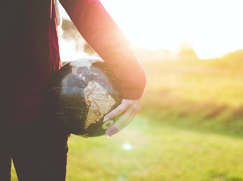 Person mit einer schwarz-braunen Weltkugel unter dem Arm, während sie auf einer Wiese zur Golden Hour steht ©by Ben White on unsplash