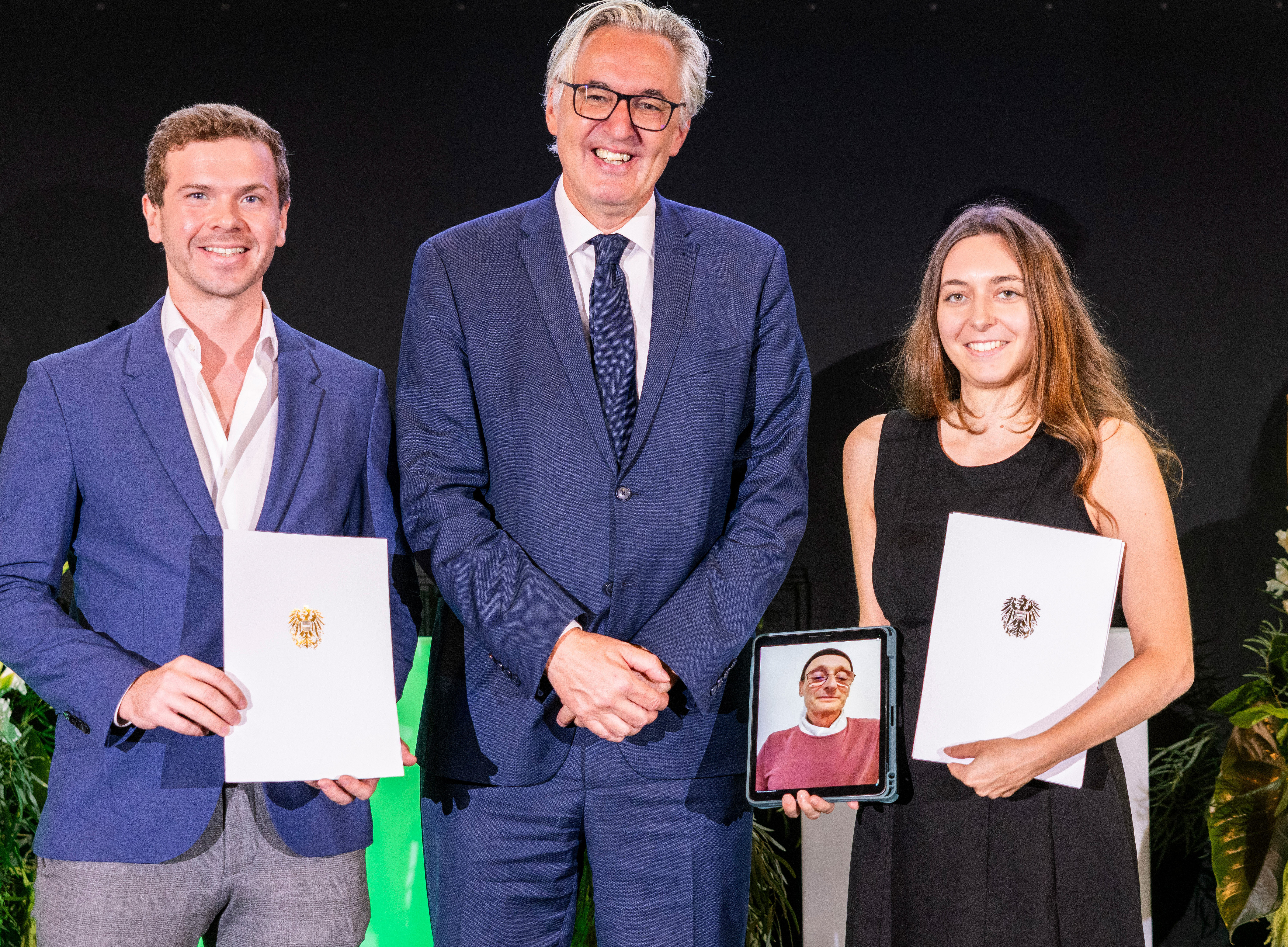 Foto von drei Personen, die bei der Zeremonie zusammen auf der Bühne stehen. Ein Mann links und eine Frau rechts halten Dokumente mit dem Vermerk „Universität“ in der Hand, festliche Atmosphäre. Ein Mann in der Mitte trägt einen blauen Anzug mit weißer Bluse und lächelt. Die Frau trägt ein schwarzes Kleid und hält ein Tablet mit dem Foto einer Person in der Hand. Der Mann links trägt eine dunkelblaue Jacke und eine graue Hose. 