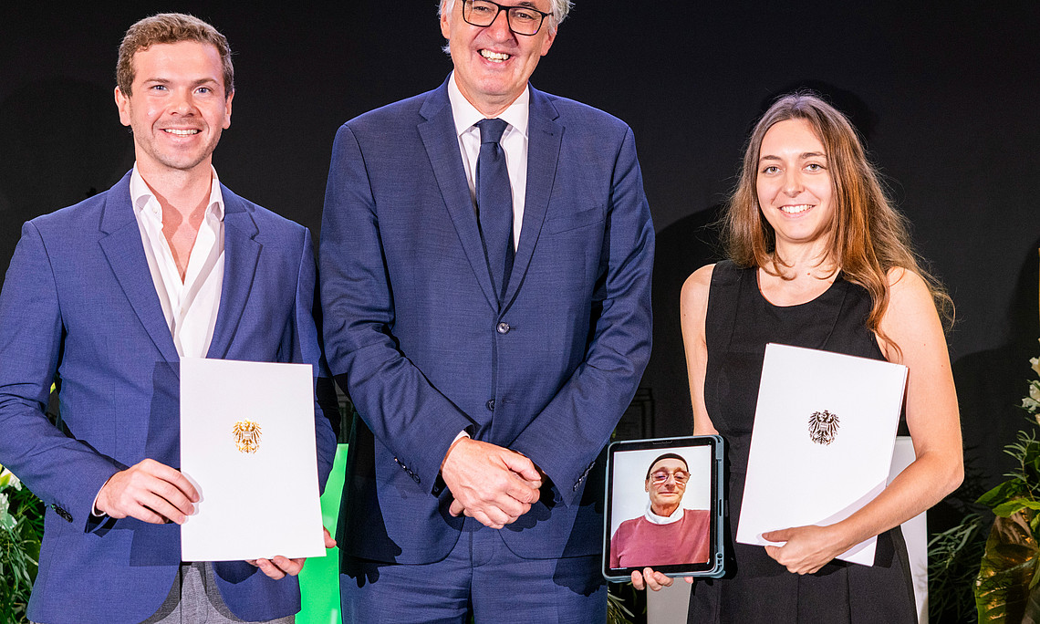 Foto von drei Personen, die bei der Zeremonie zusammen auf der Bühne stehen. Ein Mann links und eine Frau rechts halten Dokumente mit dem Vermerk „Universität“ in der Hand, festliche Atmosphäre. Ein Mann in der Mitte trägt einen blauen Anzug mit weißer Bluse und lächelt. Die Frau trägt ein schwarzes Kleid und hält ein Tablet mit dem Foto einer Person in der Hand. Der Mann links trägt eine dunkelblaue Jacke und eine graue Hose.