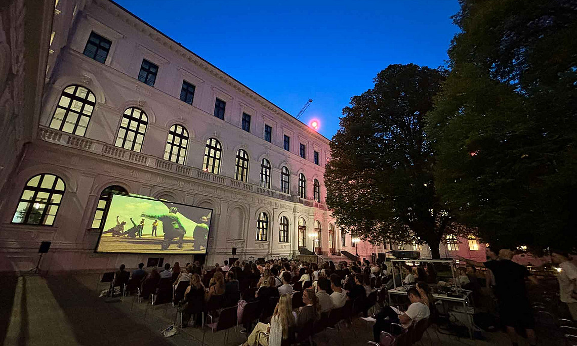 Zahlreiche Erwachsene sitzen abends auf Stühlen im Innenhof vor einer Leinwand, auf der ein Film läuft, umgeben von historischen Gebäuden und Bäumen.