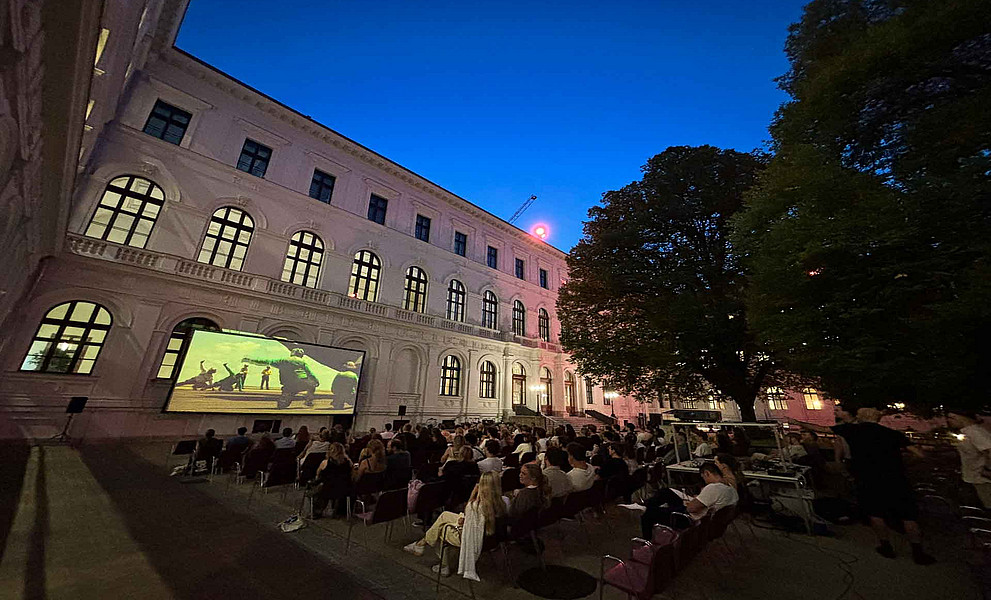Zahlreiche Erwachsene sitzen abends auf Stühlen im Innenhof vor einer Leinwand, auf der ein Film läuft, umgeben von historischen Gebäuden und Bäumen.