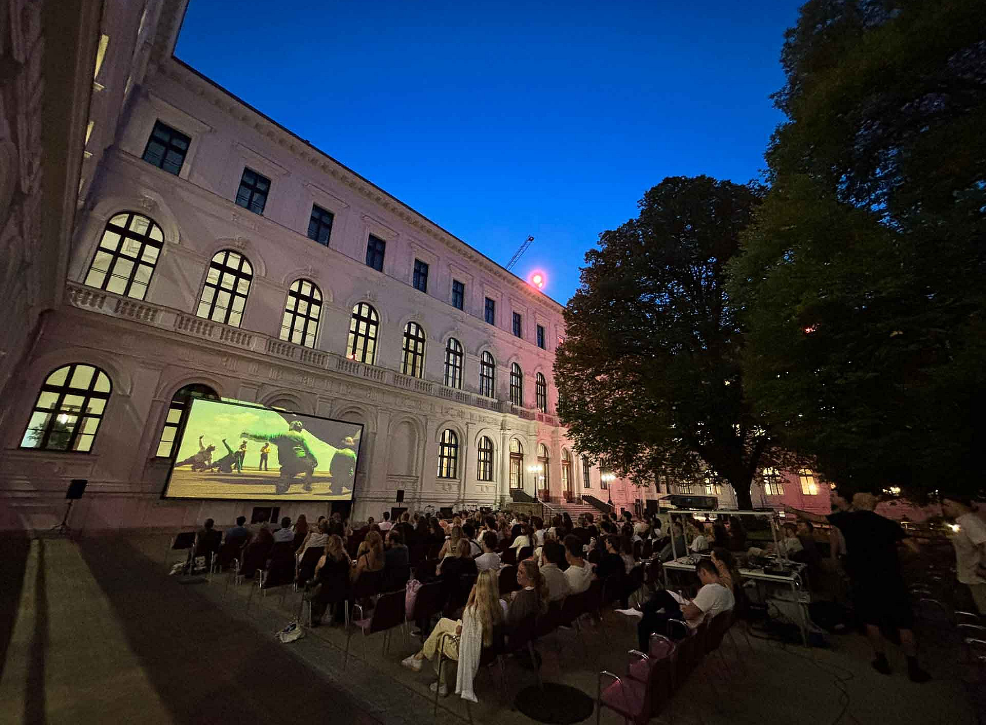 Zahlreiche Erwachsene sitzen abends auf Stühlen im Innenhof vor einer Leinwand, auf der ein Film läuft, umgeben von historischen Gebäuden und Bäumen. ©Uni Graz/Schweiger