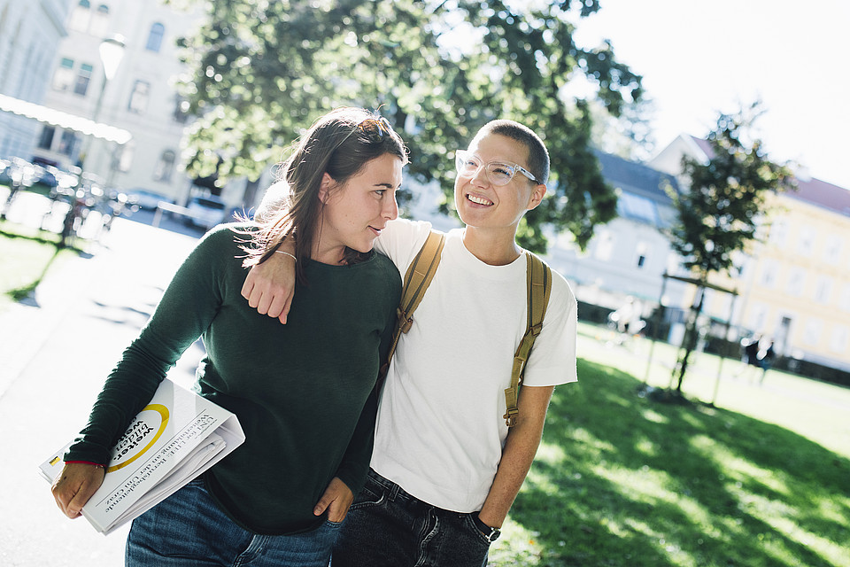 Zwei Studierende gehen gut gelaunt nebeneinander, eine hat den Arm um die andere