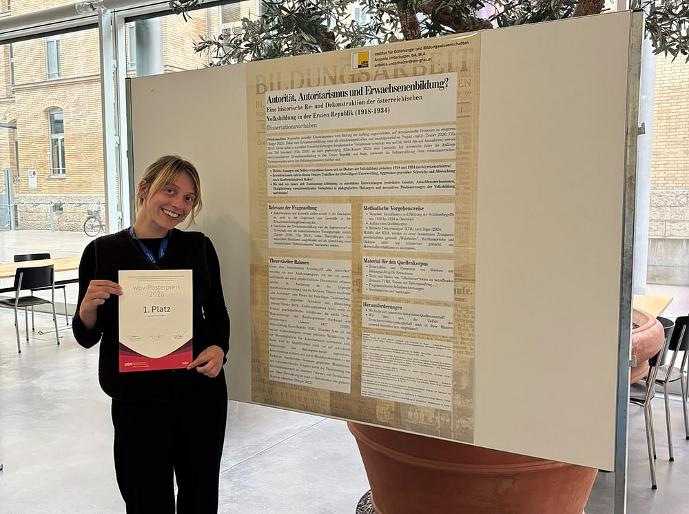 eine junge Frau, die lächelnd mit einer Urkunde neben einem Forschungsposter steht ©Uni Graz/Fachbereich Erwachsenen - und Weiterbildung