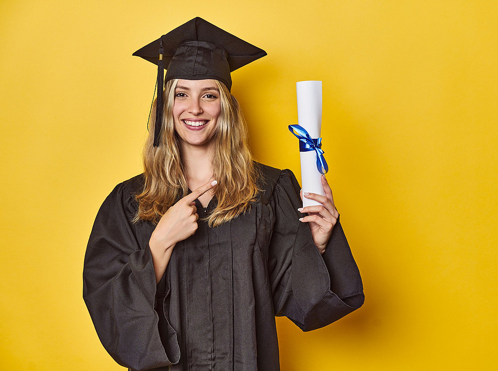 Junge Frau in Abschlussrobe vor gelbem Hintergrund hält ein Diplom in der Hand, Studienabschluss. ©Asier - stock.adobe.com