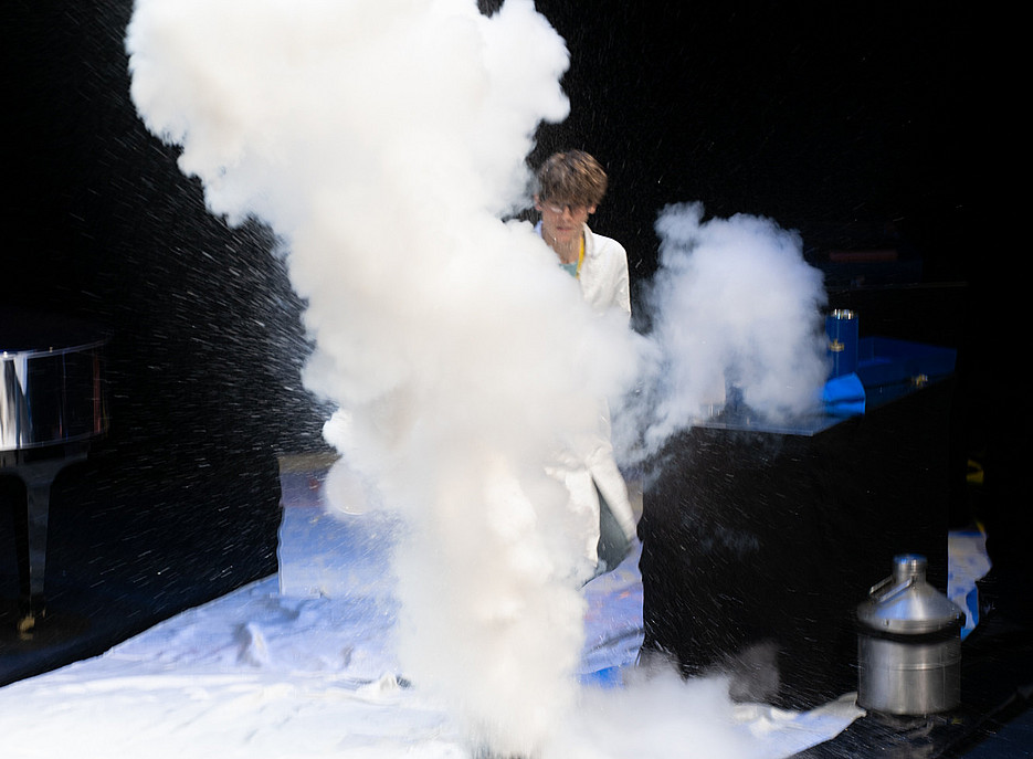 Eine Person in weißem Laborkittel steht hinter einer großen weißen Rauch- oder Dampfwolke auf einer Bühne mit schwarzem Boden und weißer Abdeckung. Links ein Flügel, rechts ein schwarzer Tisch mit einem blauen Gerät und einem silbernen Behälter. 