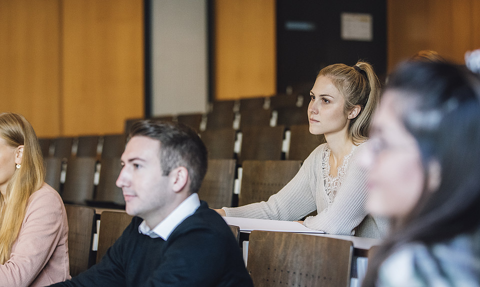 Eine Studentin sitzt mit anderen Studierenden im Hörsaal und blickt interessiert nach vorn.
