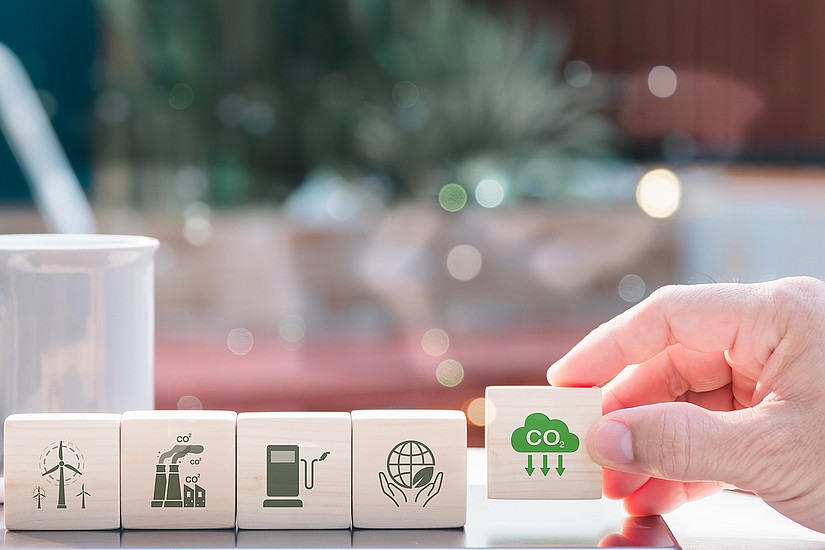 Hand lifts one of five dominoes with different symbols: Wind turbine, factory, petrol pump, globe, CO2 cloud with three arrows pointing downwards