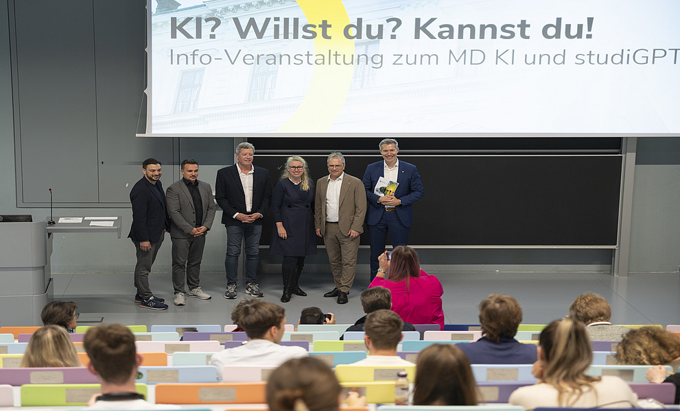 Eine Gruppe von sechs Führungskräften der Universität steht auf dem Podium vorne, auf einer großen Präsentationsleinwand steht „KI? Willst du? Kannst du?“, das Publikum sitzt in angenehmem Abstand zur Bühne.