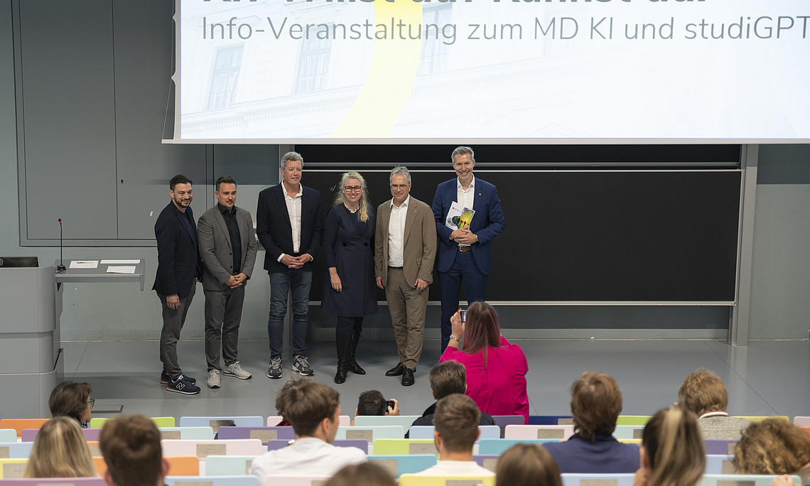 A group of six university executives stands on the podium at the front, and a large presentation screen displays the words ‘AI? Do you want it? Can you do it?’ The audience sits at a comfortable distance from the stage.