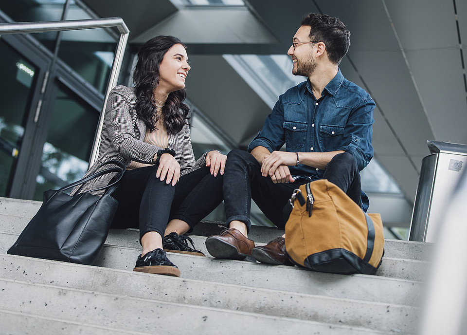 Zwei Studierende sitzen draußen nebeneinander auf den Stufen eines Universitätsgebäudes und unterhalten sich fröhlich miteinander.
