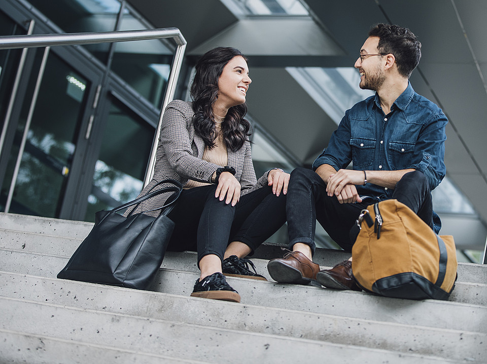 Zwei Studierende sitzen draußen nebeneinander auf den Stufen eines Universitätsgebäudes und unterhalten sich fröhlich miteinander.