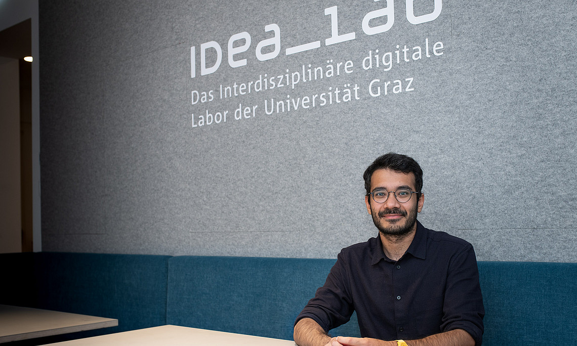 A person is sitting at a light-colored table in front of a gray wall with the inscription “IDEA_lab – The Interdisciplinary Digital Laboratory of the University of Graz.” The person is wearing a dark blue shirt and a yellow wristwatch. In the background, you can see upholstered benches in blue and part of a passageway.