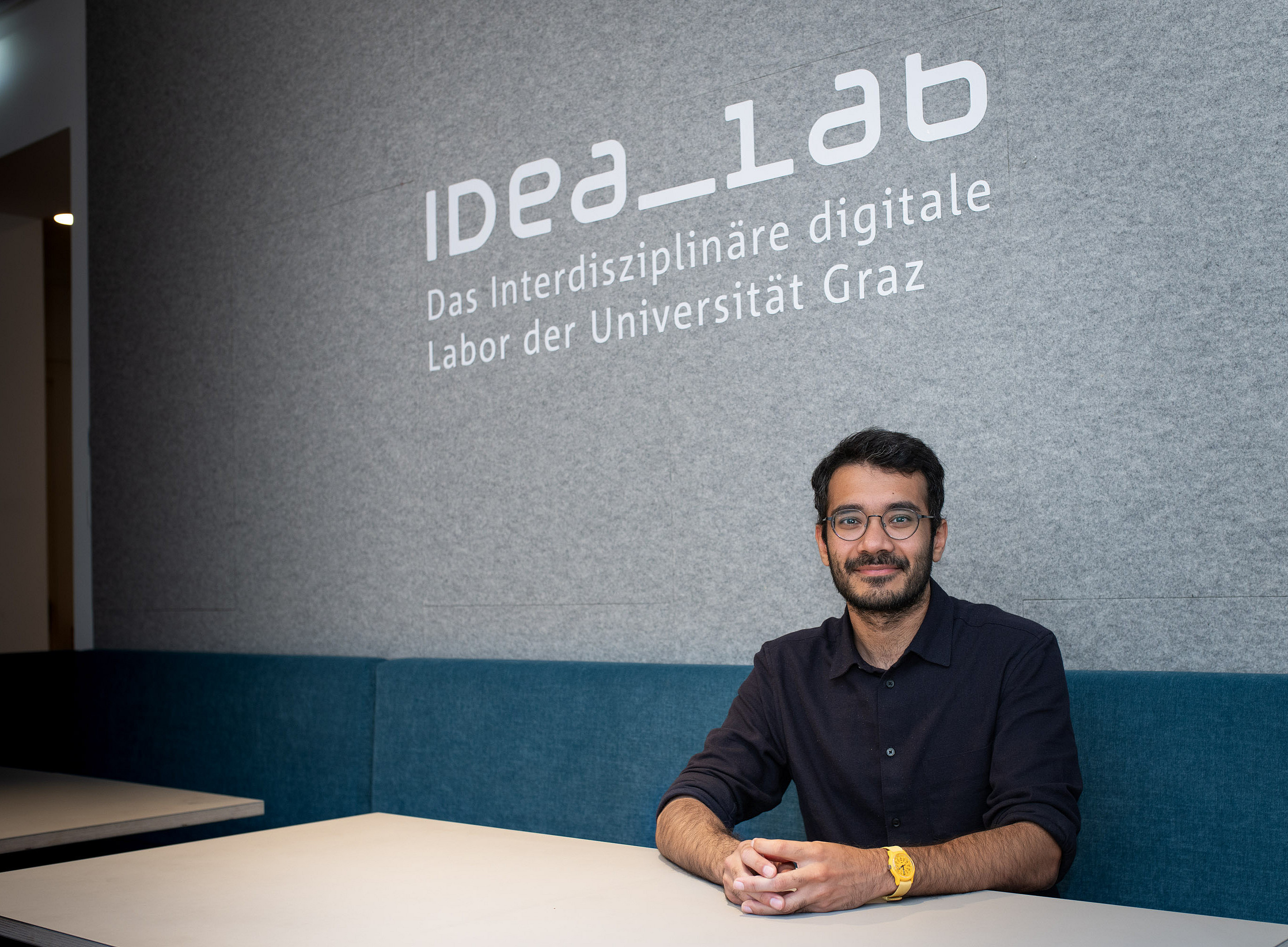 A person is sitting at a light-colored table in front of a gray wall with the inscription “IDEA_lab – The Interdisciplinary Digital Laboratory of the University of Graz.” The person is wearing a dark blue shirt and a yellow wristwatch. In the background, you can see upholstered benches in blue and part of a passageway. 