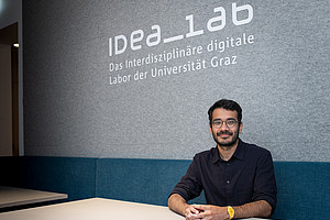 A person is sitting at a light-colored table in front of a gray wall with the inscription “IDEA_lab – The Interdisciplinary Digital Laboratory of the University of Graz.” The person is wearing a dark blue shirt and a yellow wristwatch. In the background, you can see upholstered benches in blue and part of a passageway.