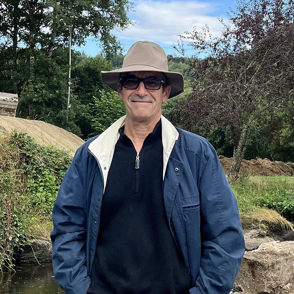 Ein Foto von Marc Bernardot, einem Mann mit Brille und dunklem Anzug, der im Freien steht, mit einer Stadtlandschaft im Hintergrund, darunter Gebäude und ein klarer Himmel. ©Marc Bernardot